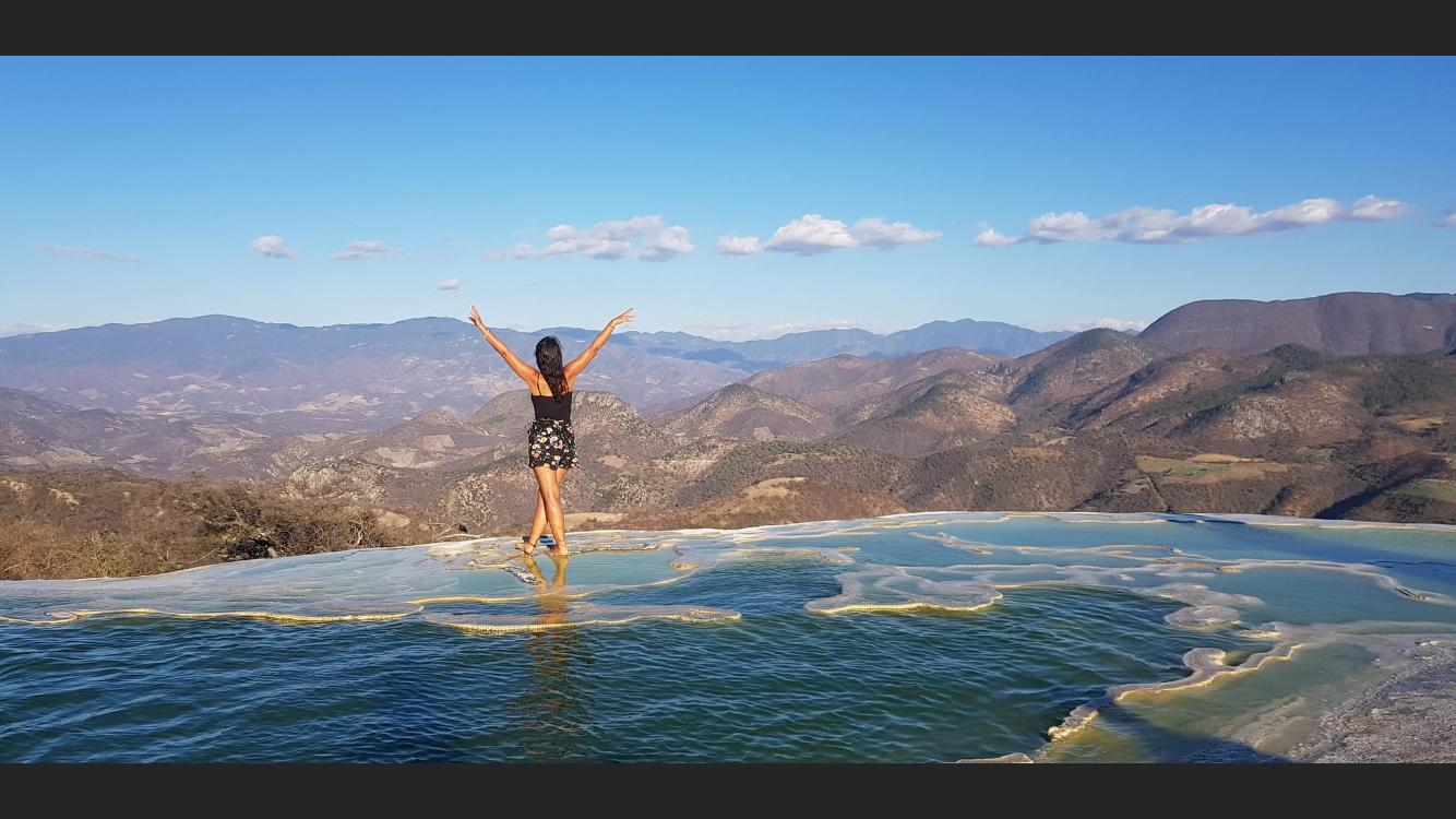 Frau mit ausgebreiteten Armen auf Steinformation mit türkisfarbenem Wasser und Bergen